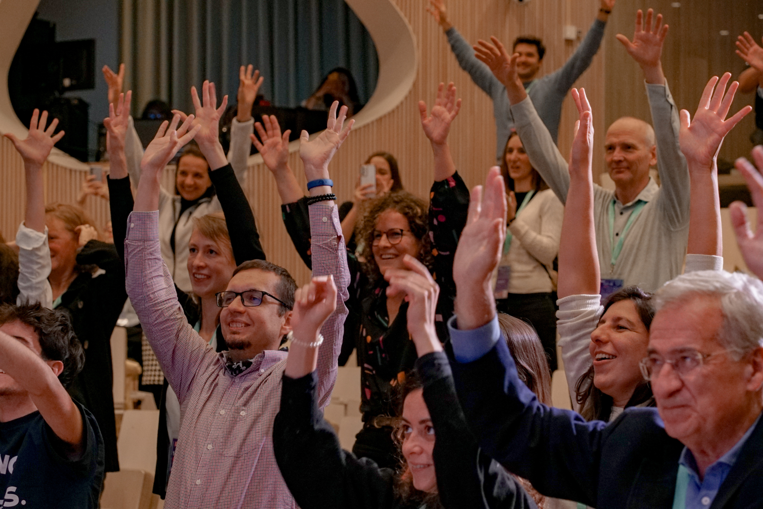 A diverse group of people enthusiastically raises their hands in a well-lit room, smiling and laughing.